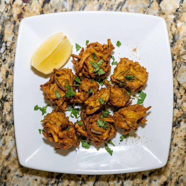 Golden-brown pakoras garnished with fresh cilantro and a lemon wedge on a white square plate.