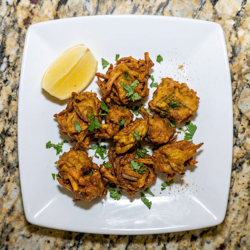 Golden-brown pakoras garnished with fresh cilantro and a lemon wedge on a white square plate.